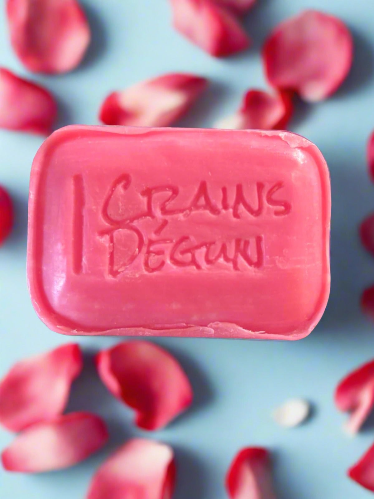 pink bar soap with crains degun engraved in the middle of the soap bar on a blue background with rose petals around the soap